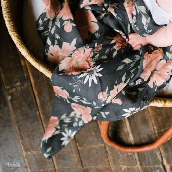 peony bloom charcoal gray swaddle blanket covering the baby bassinet with the baby hands holding a small portion of the floral swaddle blanket