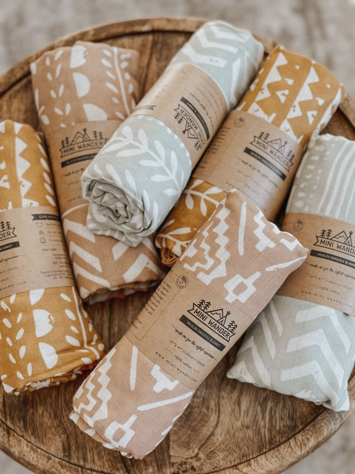 photo of our rolled boho unisex baby blanket wrapped in brown kraft paper with the mini wander logo placed on top of a wooden circular table