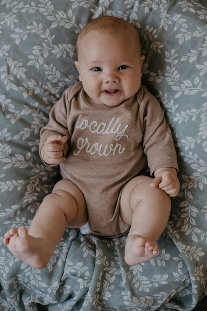 A photo of the baby in brown long sleeves onesie with a word locally grown lying down on a mini wander Leafy Sprig Blue swaddle