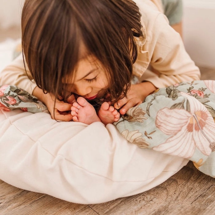 Big sister is kissing the baby's feet. The baby is sleeping on her baby nest, which is wrapped in a garden floral sea foam swaddle.