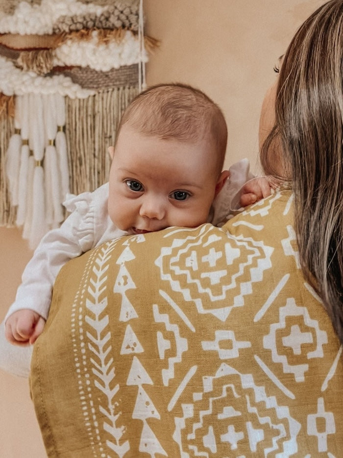 mom carrying baby wearing white clothes and peeking on her shoulders with our dark yellow blanket hanging on mommy's shoulders