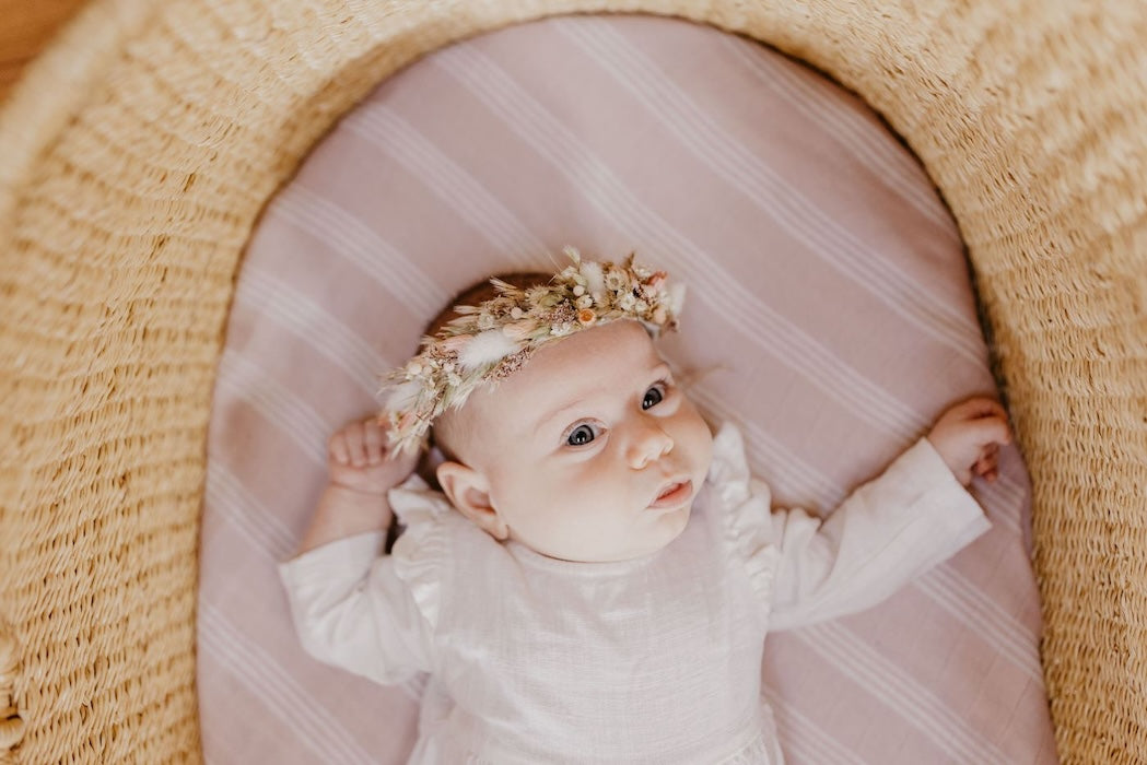 Little girl with a flower crown wearing white baby clothes laid down on her baby bassinet wrapped in a smoky rose mauve color receiving blankets