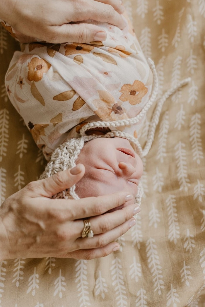 an image of a tiny little baby wrapped in sunflower swaddle and a bonnet on her head laid on top of the cream baby blanket