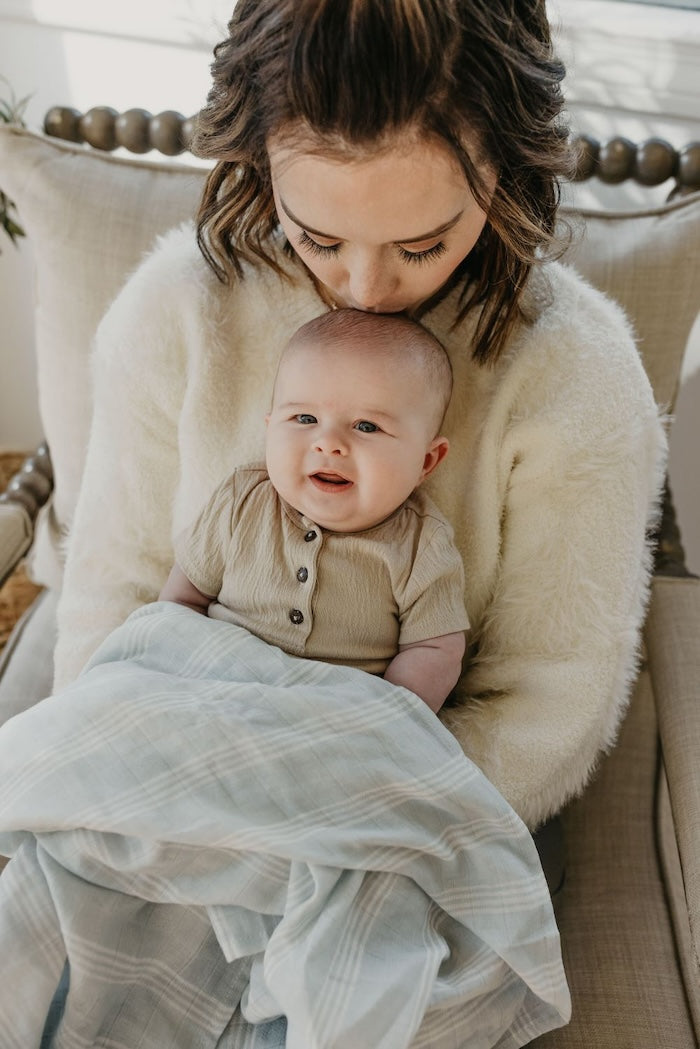 mom kissing baby's head while baby smiles at the camera and baby is wearing neutral colored clothes covered in stripe baby swaddle made with high quality cotton muslin fabric