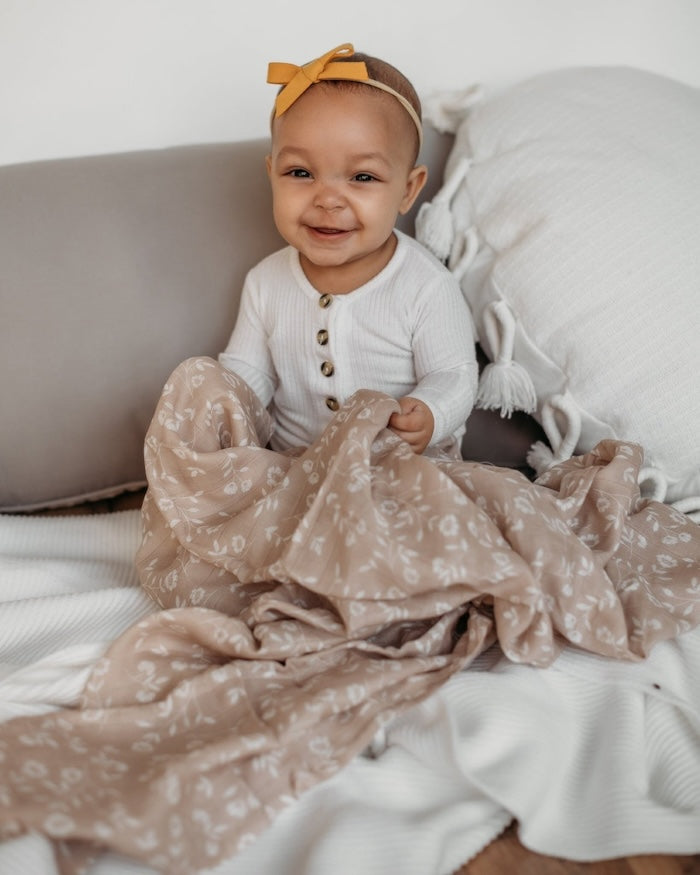 a sweet little baby holding her wildflowers taupe floral baby blanket smiling in front of the camera