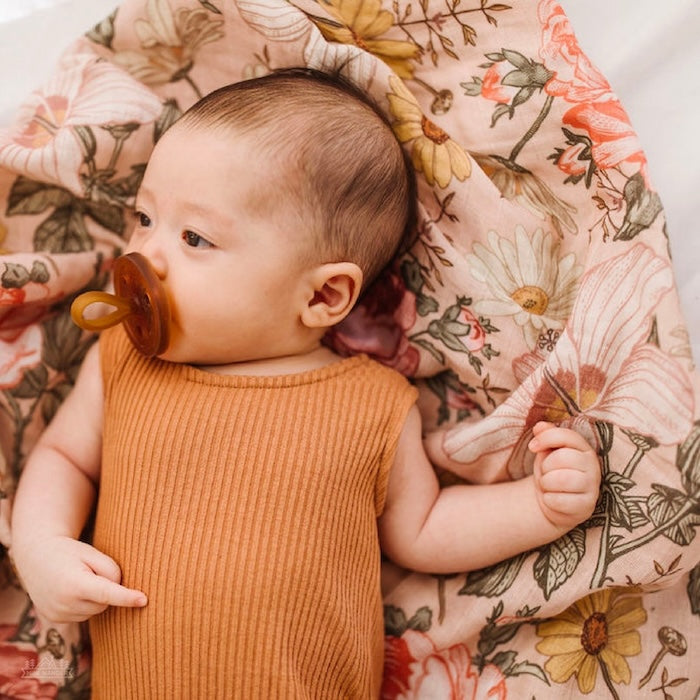 Baby in an orange onesie sucking on her pacifier, she's curled up on the garden floral swaddle, which features hibiscus, pink roses, and a yellow sunflower.