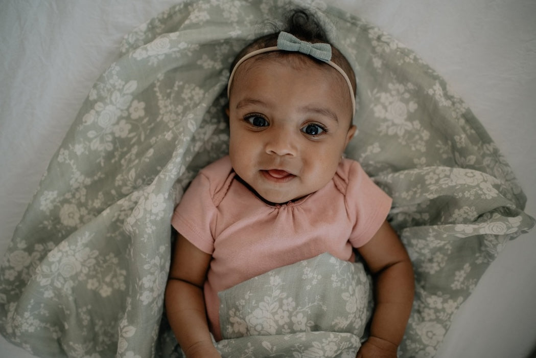 little baby girl wearing a pink top loosely wrapped in her vintage bouquet green swaddle blanket, looking happily on the camera