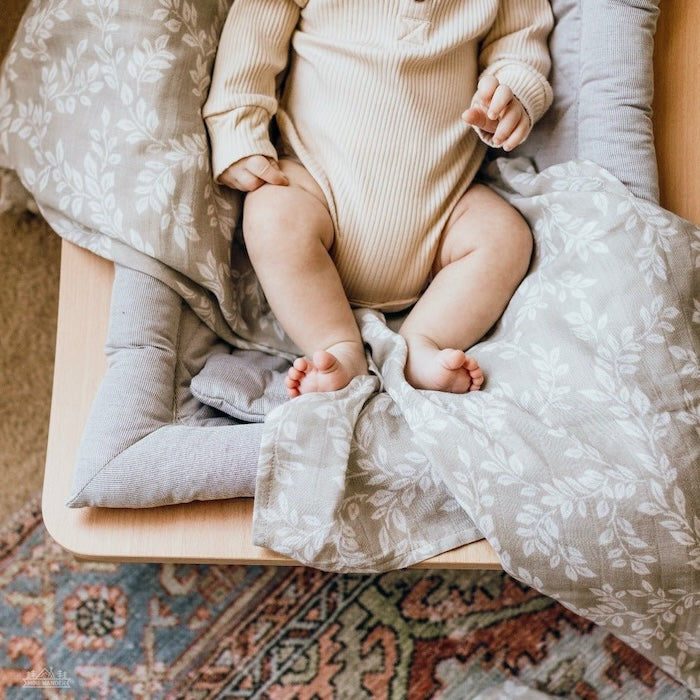 cute baby lying down in a wooden baby rocker covered by a grey swaddle with leafy design