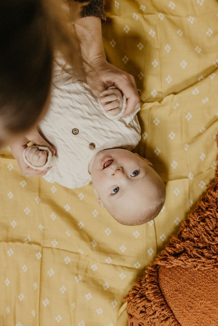 baby wearing gender neutral baby clothes lying on a yellow baby blanket holding mommy's hands while smiling
