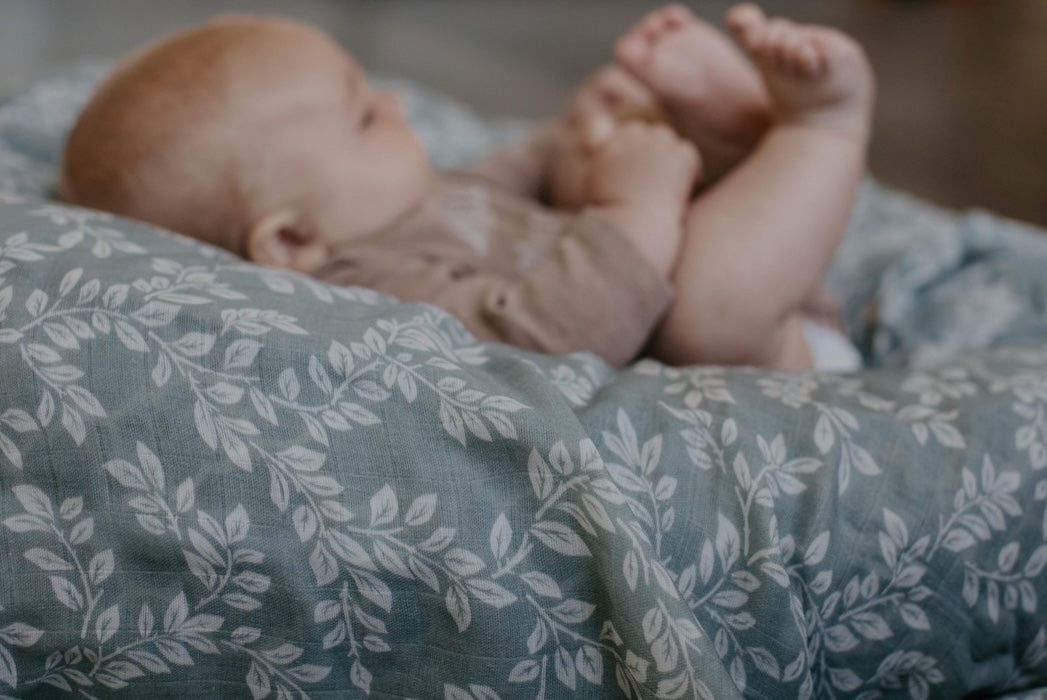 The baby is playing with his feet while lying down on a mini wander leafy Sprig Blue swaddle