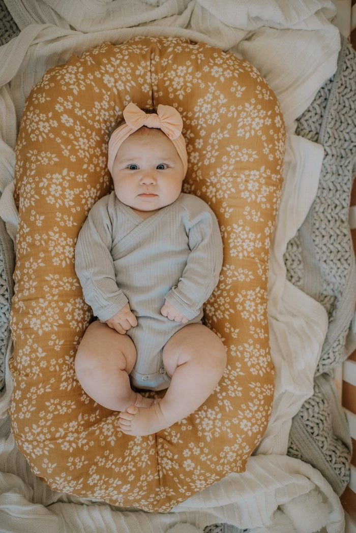 A baby with a pink bow is lying on a mustard colored Mini Wander swaddle with a white floral design.