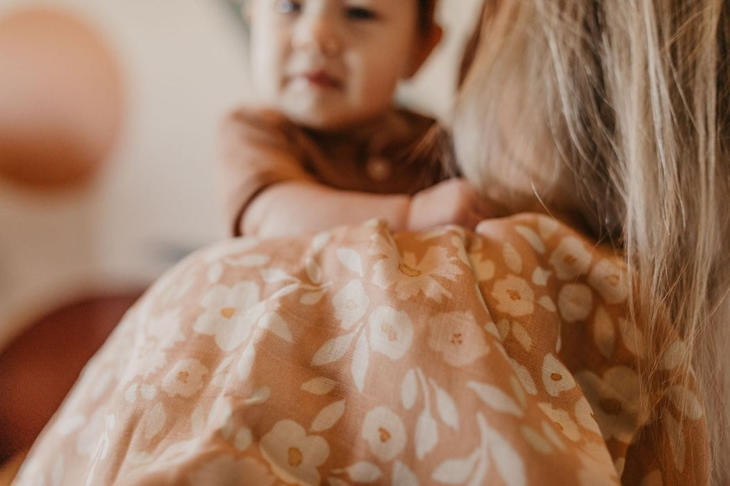 mommy carrying baby wearing brown clothes and the best baby wrap in her shoulders which is the meadow desert baby swaddle blanket, the cutest baby stuff ever