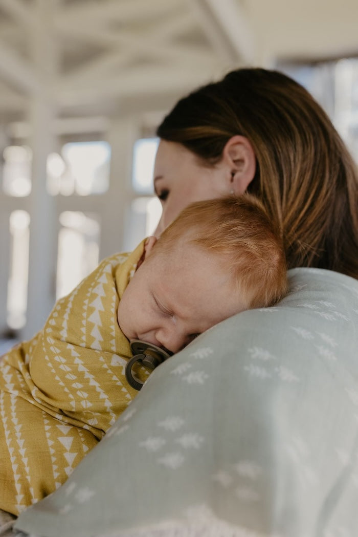little baby in a dark taupe pacifier leaning and sleeping on mommy's shoulder wrapped in our dusty yellow aztec pattern newborn blanket