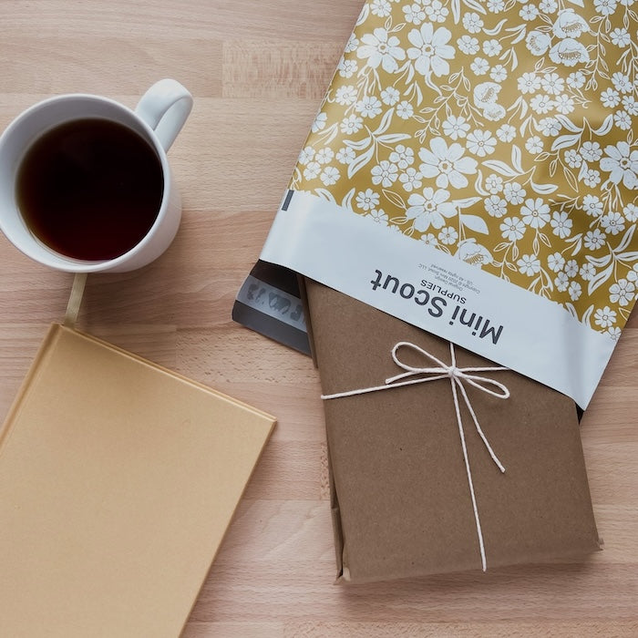 flatlay photo of the yellow 10x13 polymailer half opened with a brown package slipped inside beside a cafe americano and light brown notebook