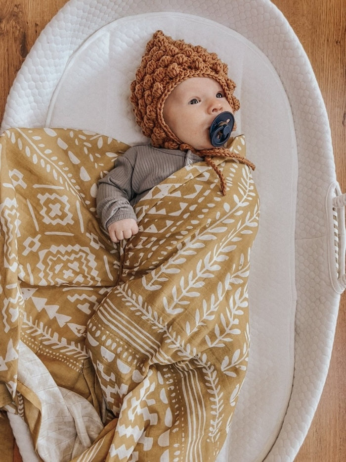 photo of a baby wearing a dark taupe clothing with a brown crochet bonnet covered and wrapped in our boho dark yellow blanket lying on a white cotton lined baby bassinet