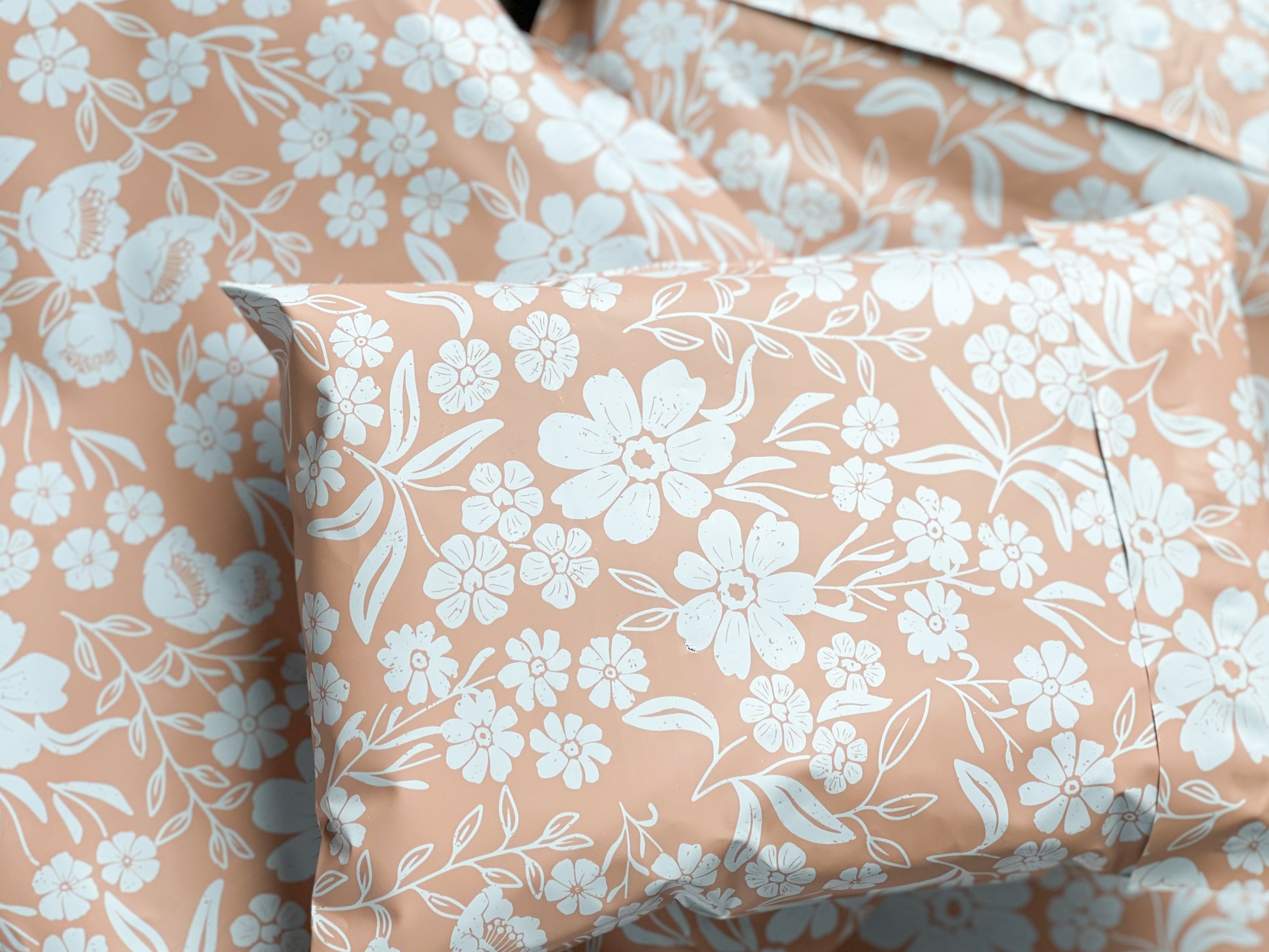 peach pink poly mailer shipping bag from mini wander. It has white flowers and a dusty pink backdrop. a small 6x9 mailing bag is on top, sealed shut with a peel and stick method.
