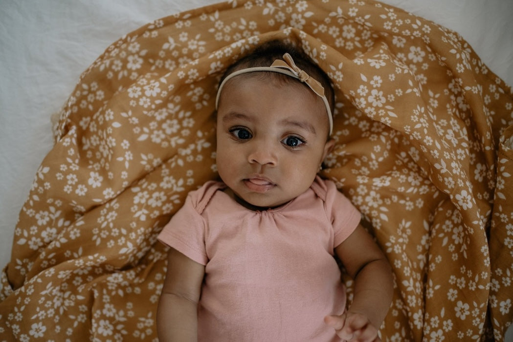 A baby wearing a brown bow is laid in a rich yellow Mini Wander swaddle with a whimsical floral design.
