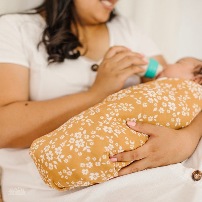 A mother feeds her baby, who is wrapped in a mustard-colored swaddle with a white floral design.