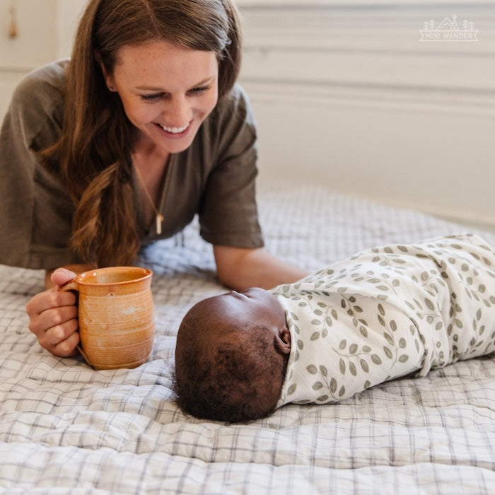 mom and baby precious moment showing mom with a mug on her hand looking happily at her sweet baby boy laying on the bed wrapped snuggly using the best summer swaddle