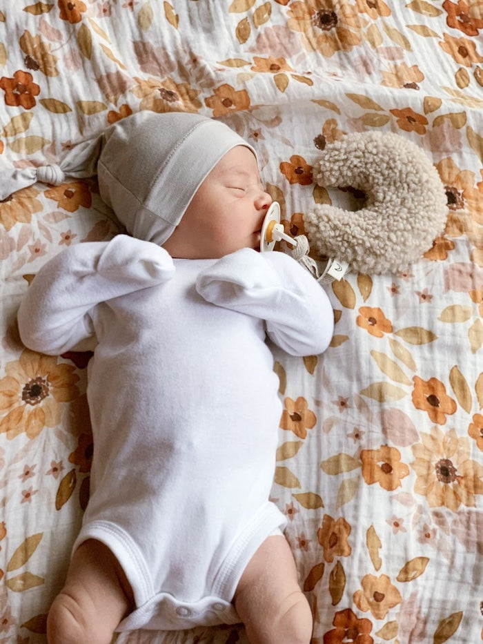 A sleeping baby wearing a bonnet and white baby clothes sleeping on top of our sunfower baby blanket beside a taupe colored moon stuffed toy