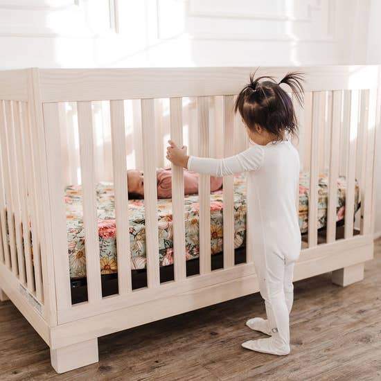 Several feet away, a young girl watches over her baby sister in a crib., who is lying on a Mini Wander cribsheet with a floral design on a green background. Flowers printed on the cribsheet include daisies, roses, and a creamy pink hibiscus.