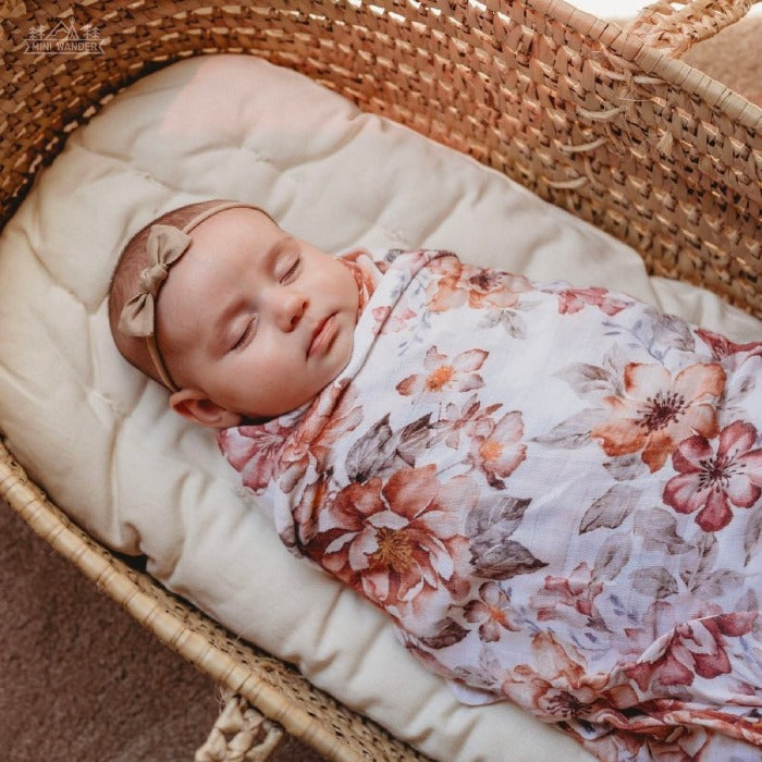 The baby is sound asleep in the baby basket. She is wrapped in a swaddle with flower pattern of fluffy primroses and ruffled peonies accented with creamy foliage from mini wander.