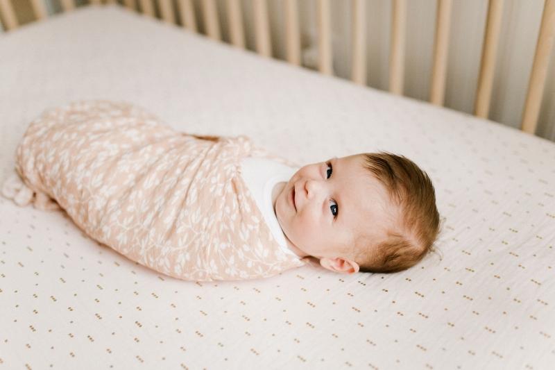 a cute smirking baby laying inside her crib wrapped like a burrito in her floral pink baby blanket