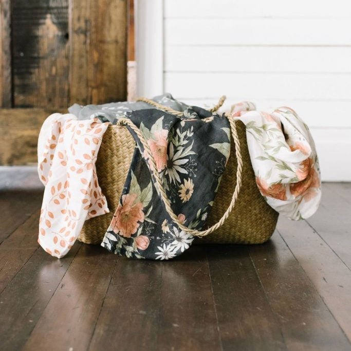 woven basket placed on the floor with three cotton swaddle tossed inside the blanket showing part of the three swaddles peeking outside the basket