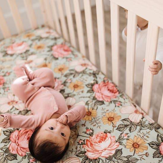 A young girl watches over her baby sister in a crib., who is lying on a Mini Wander cribsheet with a floral design on a green background. Flowers printed on the cribsheet include daisies, roses, and a creamy pink hibiscus.