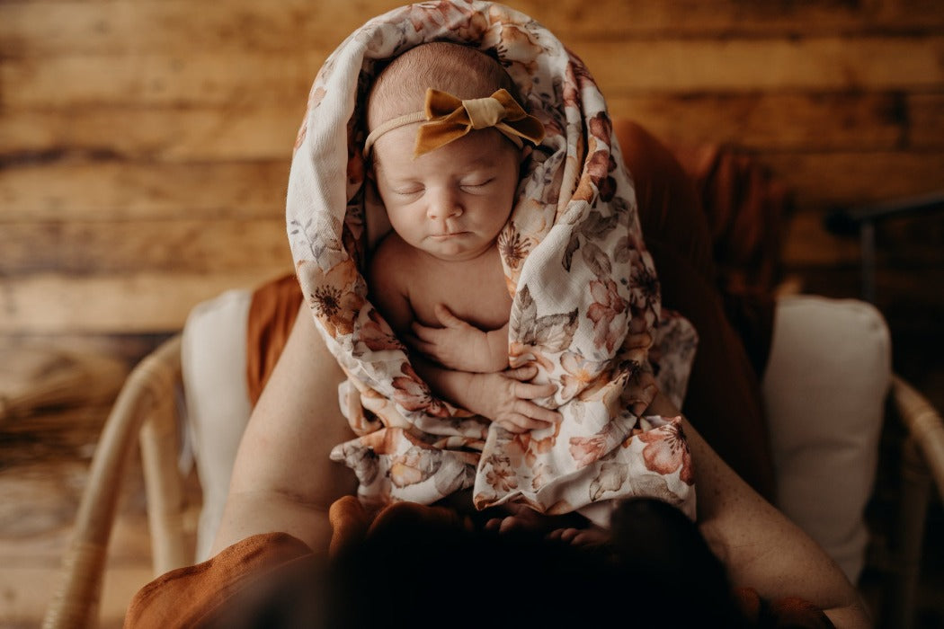 Baby in a swaddle surrounded by primroses and peonies in vintage colors. Baby is dressed cutely in a light brown bow.s wearing a cute light brown bow.