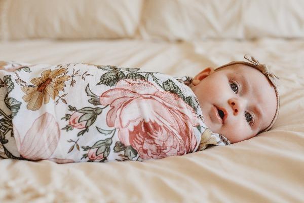 A sweet baby girl is laying down, swaddled in a wrap that has large roses, green leaves and yellow daisy flowers. The swaddle blanket is from the mini scout brand, now known as Mini Wander.