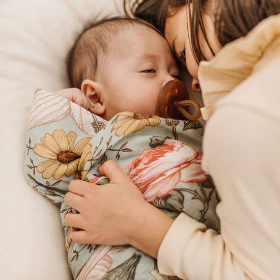 Baby swaddled in Mini Wander sea foam garden floral, napping with pacifier and receiving a hug from her sister.