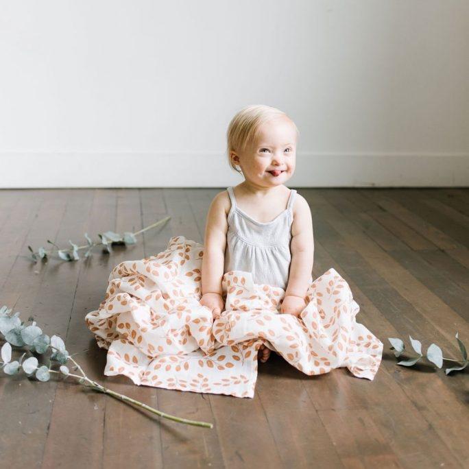 an adorable little baby girl looking at the camera with smiling with her tongue out holding the amber leaves infant summer swaddle over her lap