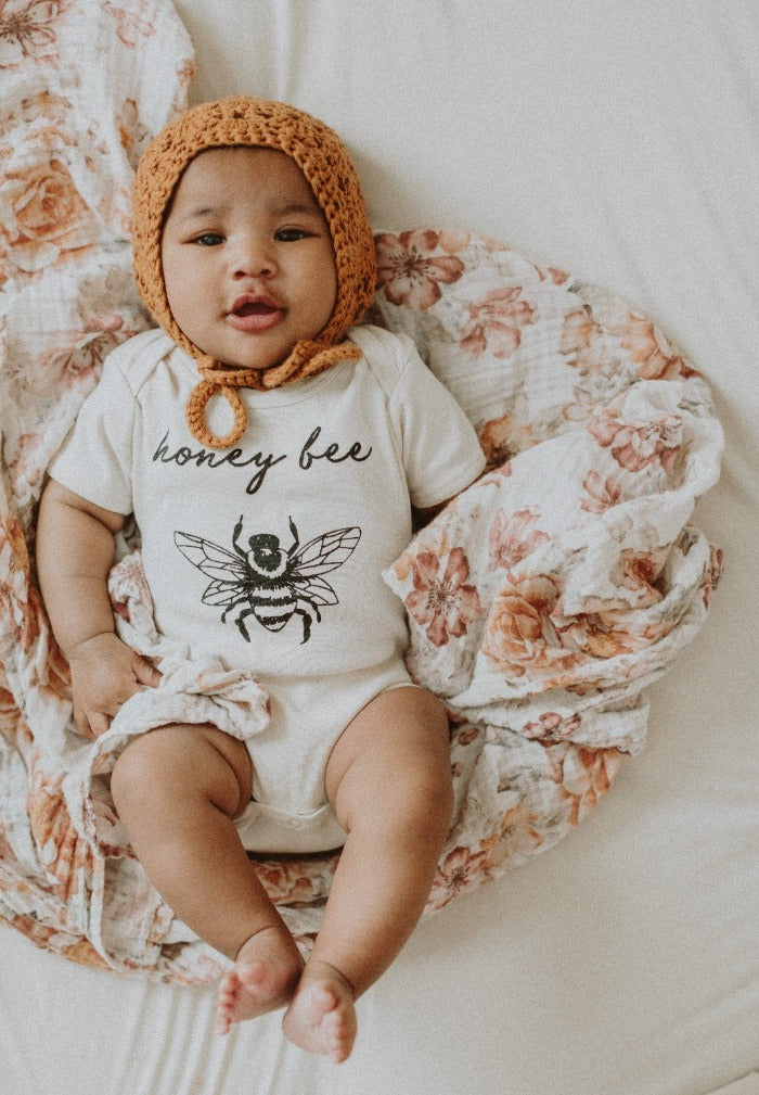 A baby with a cute orange bonnet on her head smiles at her mama while curling up on a sunset colored floral swaddle.