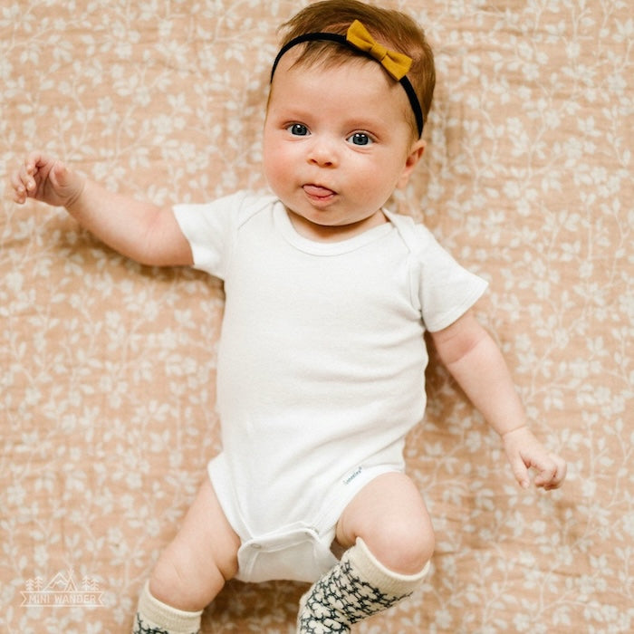 an adorable baby girl wearing a white baby romper and mustard headband and knee-socks laying on top of her beautiful pink swaddle blanket used as a photo background