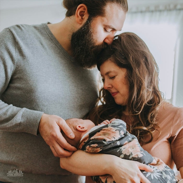 mom and dad sweet moments with the little one wrapped in our peony bloom charcoal gray blanket. dad is kissing the mom's head and mom is holding the baby in her arms