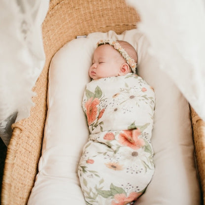 relaxing little moment of a baby girl wearing a flower crown headband swaddled in the Spring Blossom white peony swaddle lying in her baby lounger placed inside a bassinet