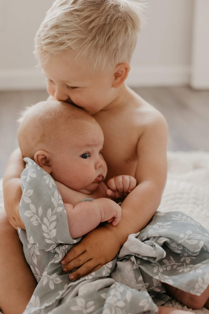Photo of a big brother kissing the forhead of his baby brother who is wrapped in a swaddle with sprouting twig design