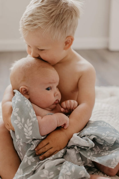 Photo of a big brother kissing the forhead of his baby brother who is wrapped in a swaddle with sprouting twig design