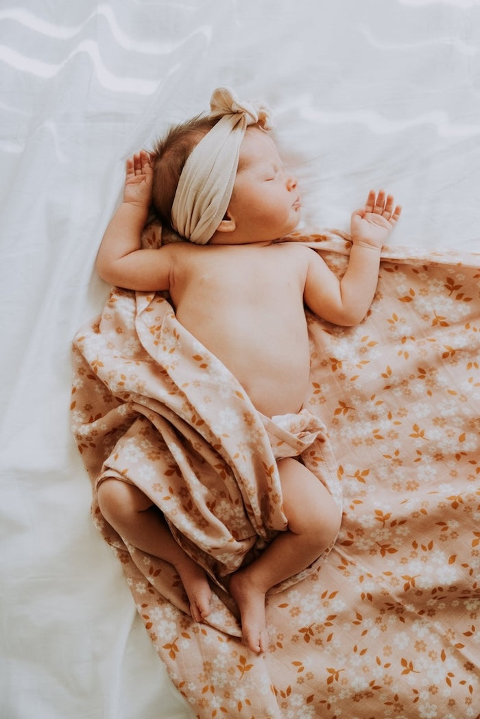 A baby with a bandana is sleeping on a loosely wrapped peach floral daisy swaddle.