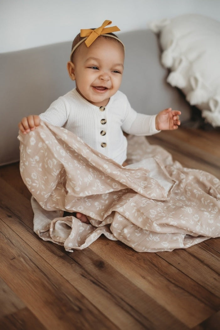 a charming baby girl sitting on a wooden floor, smiling while holding the wildflowers taupe floral baby blanket in her one hand