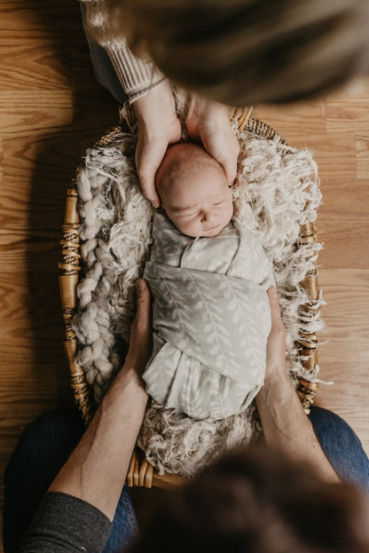 photo of a newborn baby wrapped in our tapestry boy swaddle held with love by his mom and dad while trying to lay him down in the baby bassinet