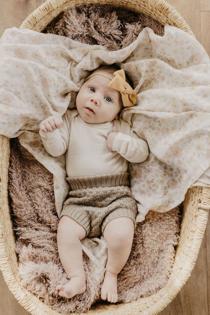 baby girl in a baby bassinet covered with meadow cream swaddle in natural cotton wearing neutral-colored clothes and yellow headband