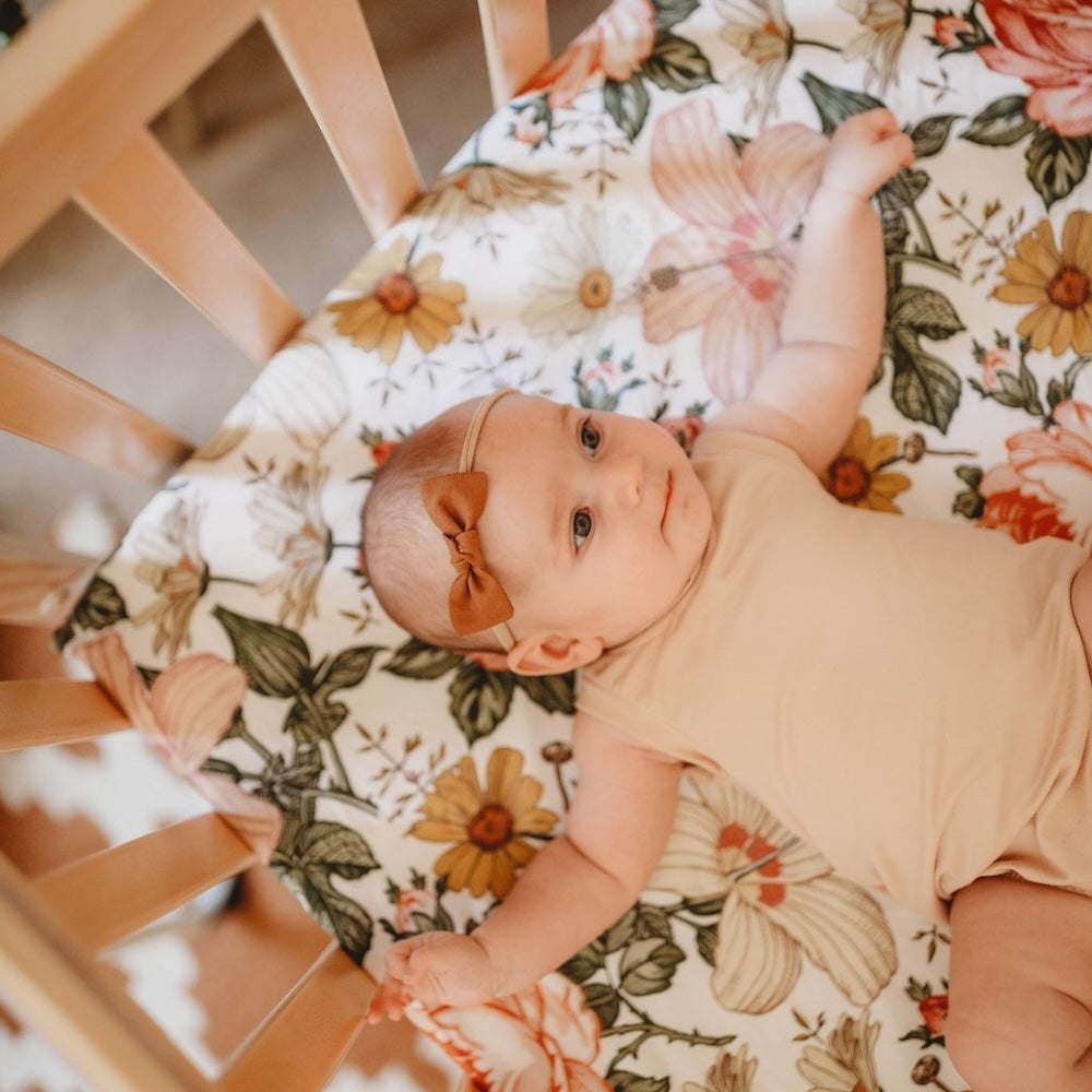 sweet darling baby wearing a brown ribbon headband and neutral baby clothes lying in her back on the mattress covered wiht the beautiful Mini Wander floral crib sheet
