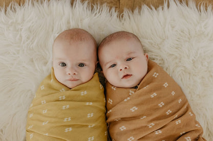 twin babies in a swaddling muslin blanket in a brown and yellow baby blanket innocently looking at the camera smiling