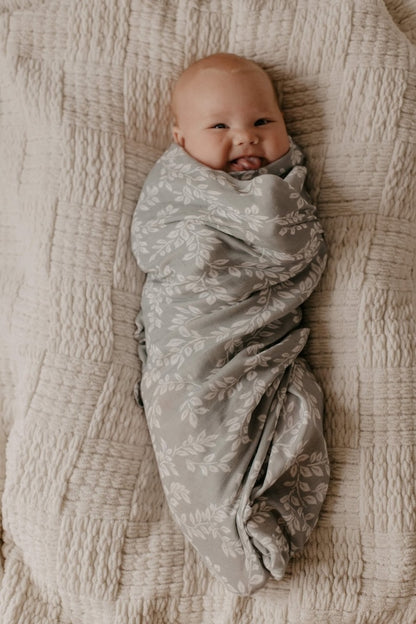A baby making funny faces and showing his tongue to the camera while wrapped in a blue swaddle decorated with twig leaves and lying down on a cream colored blanket.