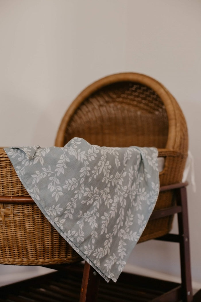 A swaddle with a leafy sprig design hangs in a native rattan baby basket.