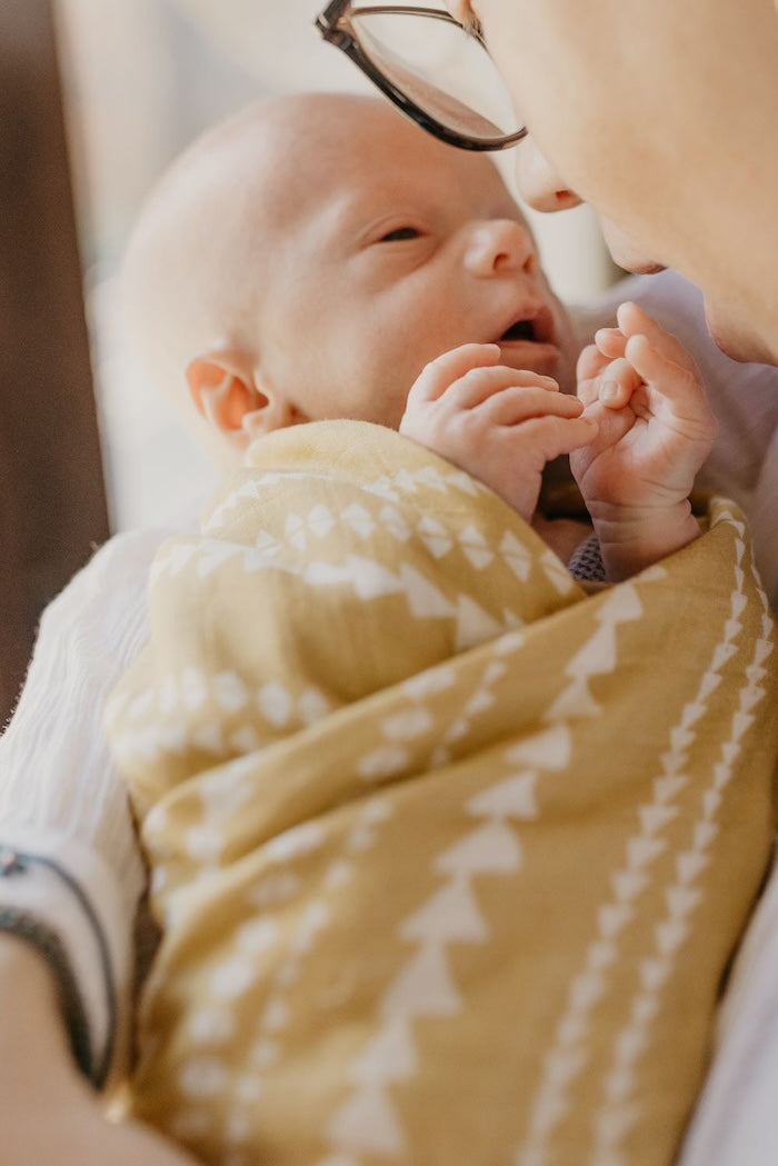 an image of mom and baby in which baby is wrapped around mommy's arms in our dusty yellow aztec pattern blanket whispering and talking to baby while baby is trying to get some restful sleep