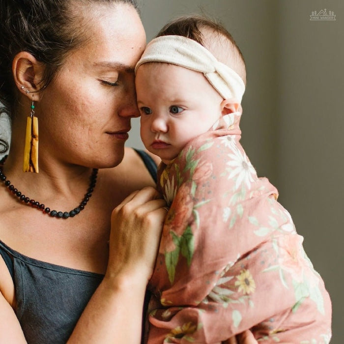 a photo of mom and daughter sweet moment in their boho nursery showing the mom carrying her swaddled baby wearing our spring blossom muslin baby swaddles