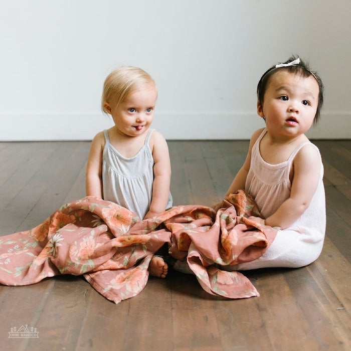 two adorable baby girls sitting on the wooden floor with the boho nursery with flower design clay brown swaddle blanket
